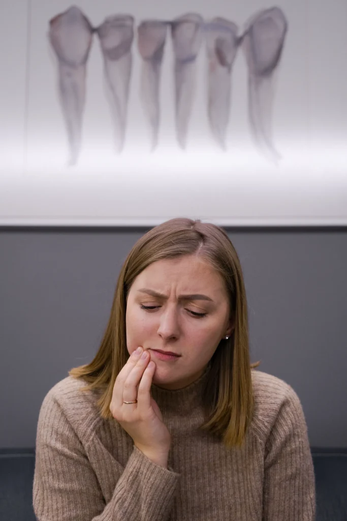 Home - Clínica Dental Dentinos Mujer sentada en una clínica dental tocándose la mandíbula con expresión de dolor.