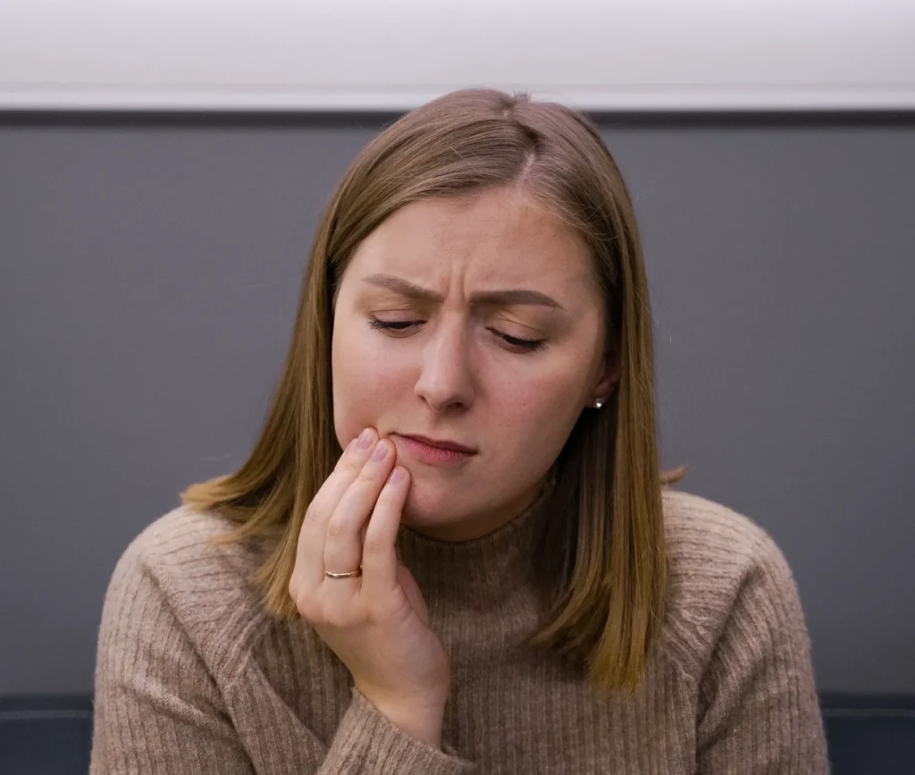Home - Clínica Dental Dentinos Mujer sentada en una clínica dental tocándose la mandíbula con expresión de dolor.