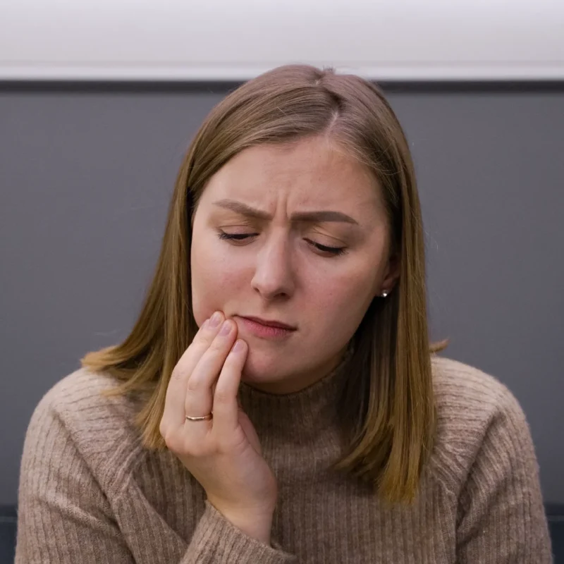 Mujer sentada en una clínica dental tocándose la mandíbula con expresión de dolor.