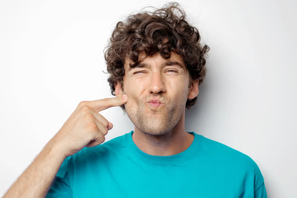 Home - Clínica Dental Dentinos Hombre señalando su mejilla y mandíbula con expresión de incomodidad o dolor muscular facial.
